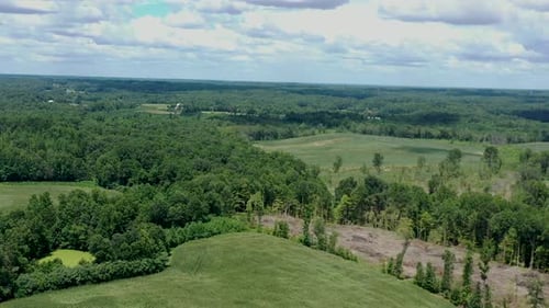 Drone flyover of a cornfield and rural wooded land with vibrant cloudy skies in the background.