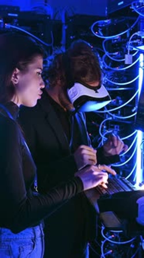 Woman programming in a server room while a man is using a Virtual Reality headset. Vertical