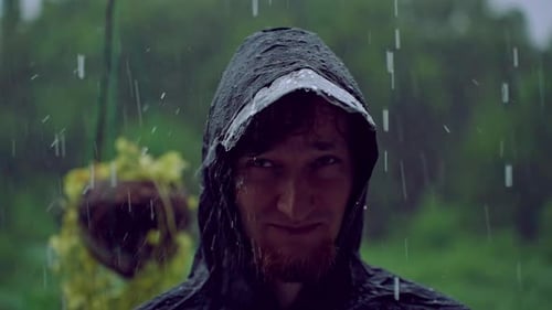 Ein Mann steht unter starkem tropischem Regen in einem dunklen Regenmantel mit Kapuze, während Wasser über sein Gesicht fließt