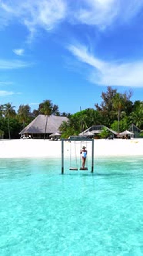 Woman Swings Above the Crystal Clear Sea on a Wooden Swing Facing the Beach and Lush Tropical