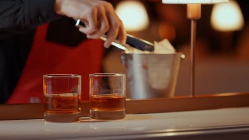 Closeup Bartender Hands Putting Ice Cubes with Forceps in Glasses in Bar