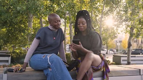 Women Friends Using Smart Phone In Sunny Urban Park