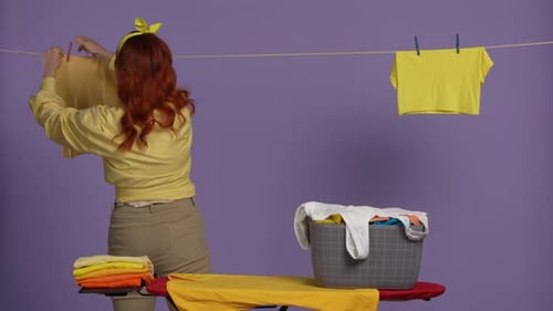 Woman in Casual Clothing Standing Next to Ironing Board with Basket Full of Laundry and Hanging the