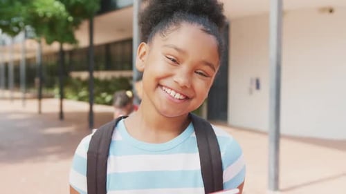 Video portrait of smiling biracial schoolgirl holding her books outside school