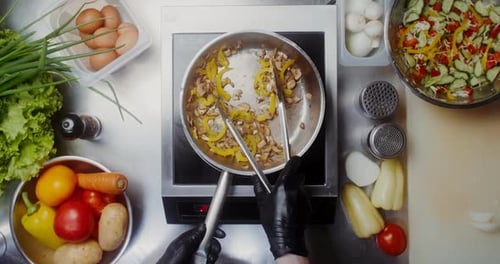 A woman in disposable black gloves mixes mushrooms with pepper in a frying pan with tongs, top view,