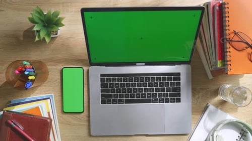Ansicht von oben nach unten auf ein Smartphone und einen Laptop mit grünem Bildschirm auf einem Holzschreibtisch