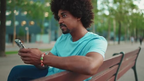man in blue t-shirt sitting on park bench using his smartphone