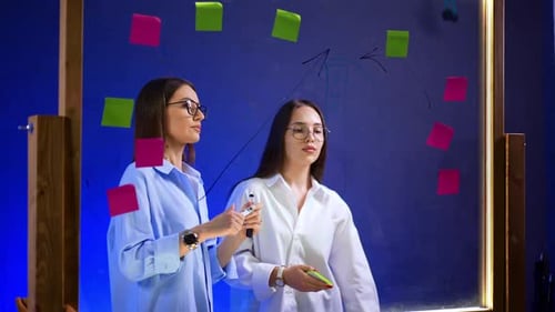 Busy ladies are standing at the glass board in the office. Colleagues draw graphs and put stickers o