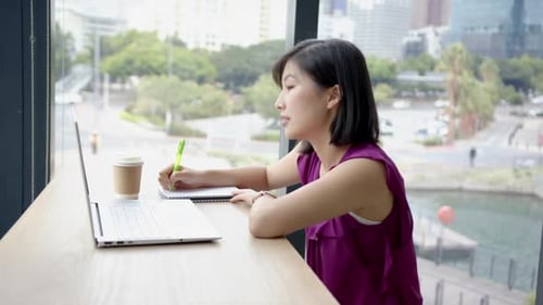 Writing in notebook, woman working on laptop with coffee cup in office