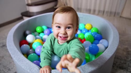Joyful happy toddler smiling broadly stands in soft basin filled with colorful balls.