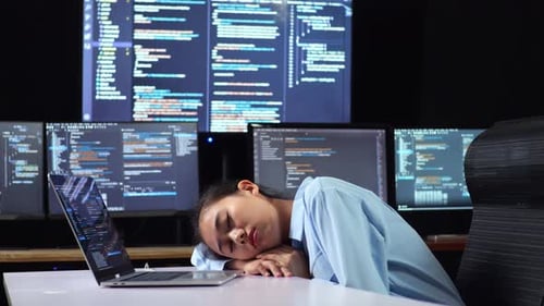 Vue latérale d'une programmeuse asiatique endormie pendant qu'elle écrit du code sur un ordinateur portable utilisant plusieurs moniteurs