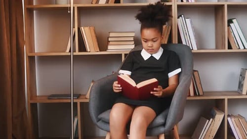 Sitting in the chair and reading the book. Cute african american girl in school uniform is at home