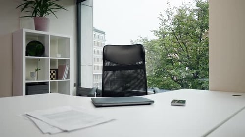 Im modernen Büro mit einem großen Fenster neben einem weißen Tisch mit einem Laptop befindet sich ein schwarzer Bürostuhl