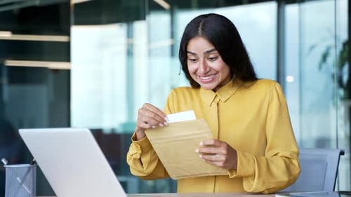 Happy young businesswoman sitting at desk in modern office reading letter with great news. Smiling