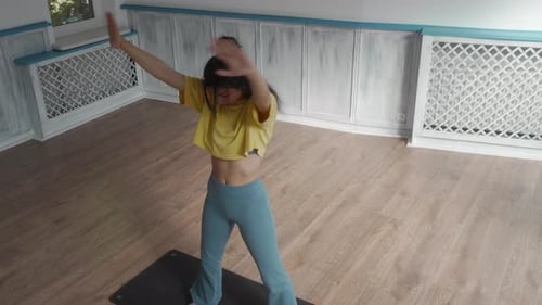A woman stretches her arms and body while standing on a yoga mat in a studio.