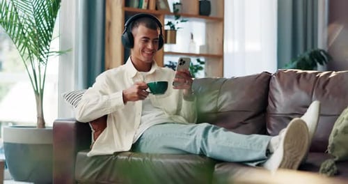 Phone, coffee and man with headphones on sofa in morning, watching or funny online video in home