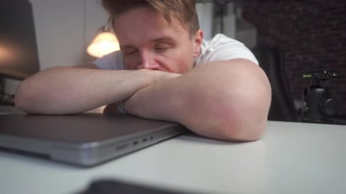 A young man with dark blonde hair leans over his desk and computer completely exhausted before he ge
