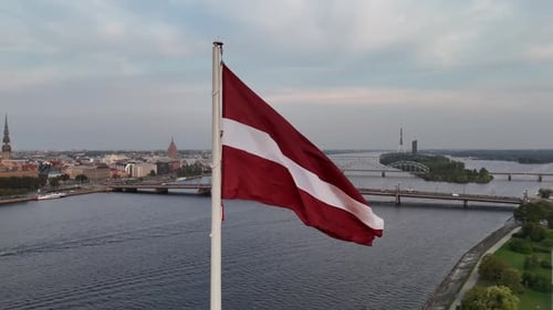 Luftaufnahme der lettischen Flagge, die am Fahnenmast in der Hauptstadt Riga bei Sonnenuntergang weht Daugava River Slow