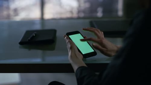 Over the Shoulder View of Businesswoman Sitting at Her Desk and Using Smartphone with Green Screen.