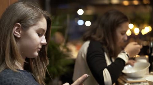 Two Teenage Girlfriends Talking Over Smartphone Sitting In Cafe