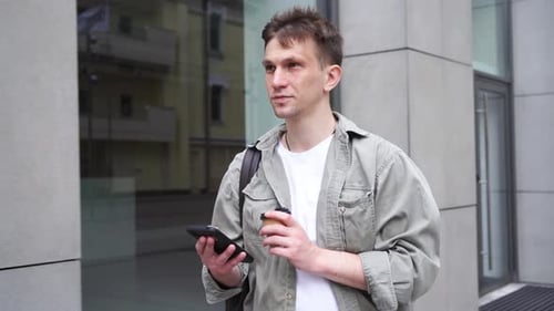 A young male entrepreneur strolls through the metropolis with a smartphone and a cup of coffee.