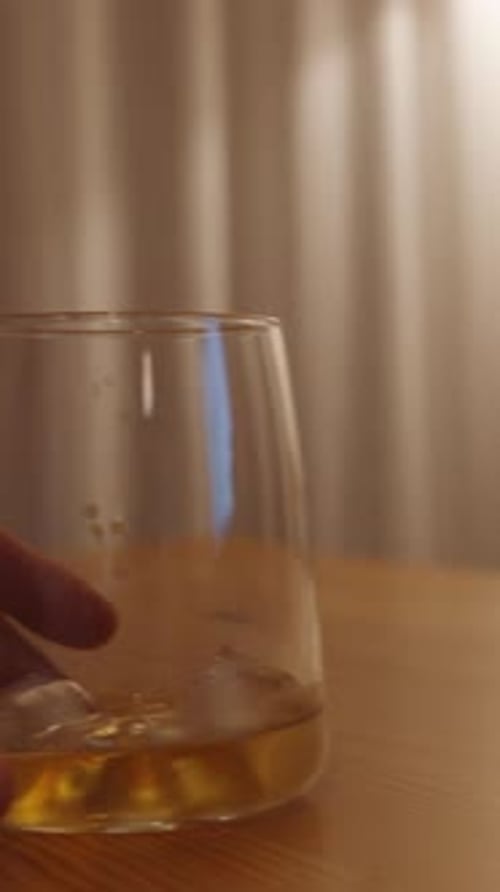 Vertical video. The man moves a glass of whiskey with ice on a wooden table. Warm light from a lamp