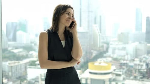 Elegant young businesswoman talking on cellphone by office window with city view