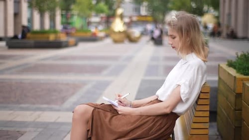 Side View of Attractive Focused Female Artist Painting in Sketchbook with Pencil Sitting on Bench on
