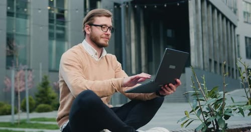 Man working or studying using laptop while sitting crossing legs on bench of city financial district