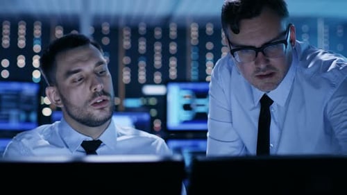 Two Professional Technicians Discussing Problems Shown on Multiple Monitors in a System Control Cen
