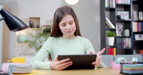 Cute Caucasian Teen Girl Sitting at Desk in Cozy Living Room and Playing on Tablet Device At Home