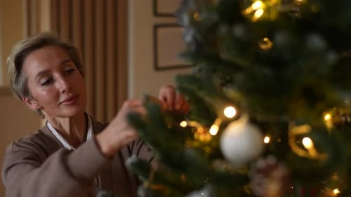 Tracking Shot of Happy Middleaged Woman Hanging Up Ball on Branch of Christmas Tree in Cozy Room