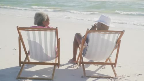 Happy senior african american couple sitting on deck chairs at beach, in slow motion