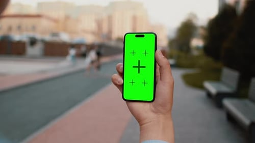 Closeup of a Hand Holding a Smartphone with a Mockup Green Screen with a Sidewalk of a City Street