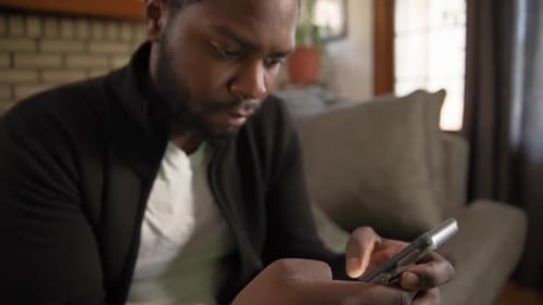 Black man texting on phone concentrates in living room close up tilt