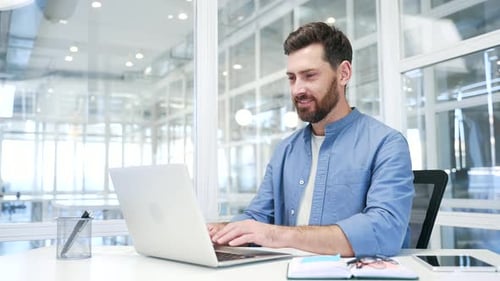 A smiling businessman working on a laptop sitting at a workplace in business office. Handsome bearde