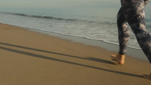 Eine junge Frau läuft barfuß und in Leggings am Strand entlang am Meer entlang, Nahaufnahme, Zeitlupe. Lo