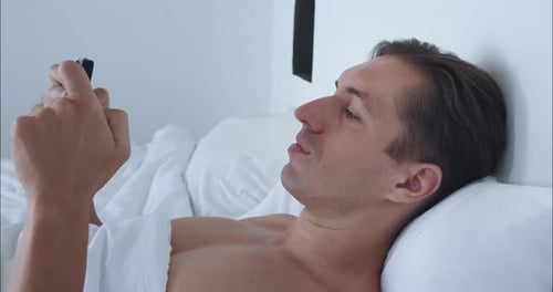 Millennial Sits Comfortably in Bed and Enjoys Playing on Phone In Bed Man is Completely Immersed in