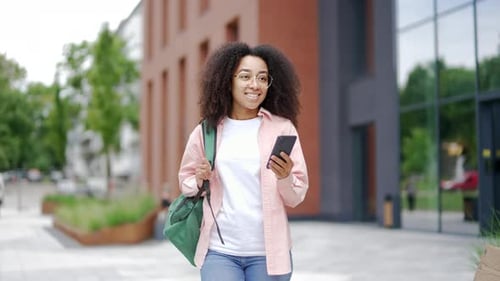 Smiling african american female student using phone walking on campus space near university building