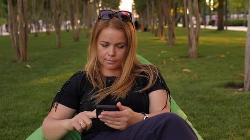 A Woman Sits in an Urban Modern Park in a Soft Chair with a Phone in Her Hands
