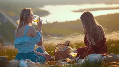 Two Blonde Women Sitting on the Field and Having a Picnic Enjoying a View of the River on Sunset