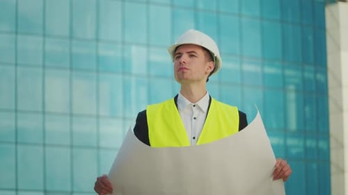 Male Engineer Wearing in Helmet and Safety Jacket Looking Construction Drawings in His Hand