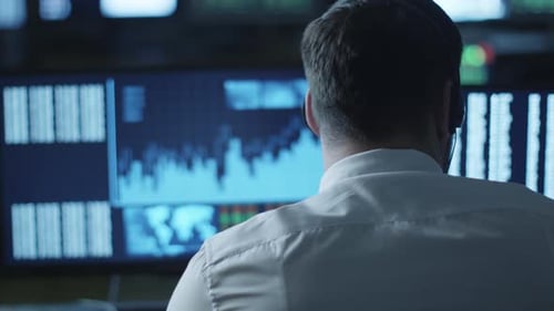 Stockbroker is talking on a headset while working on a computer in a dark office filled with displa