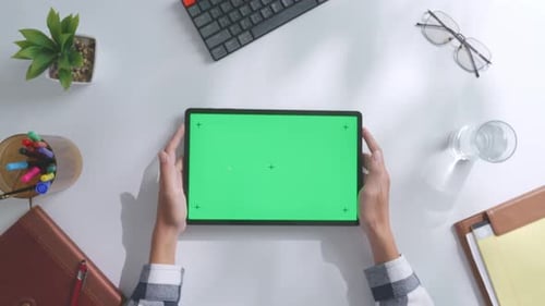 Top down view of hands holding a mock up green screen tablet on a white desk