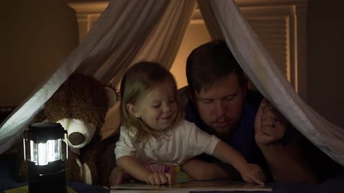 Father reading to toddler daughter at bedtime in blanket fort