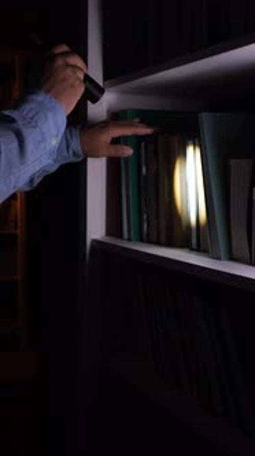 Vertical Video Man Searching for Book on Bookshelf Using Flashlight in Darkness
