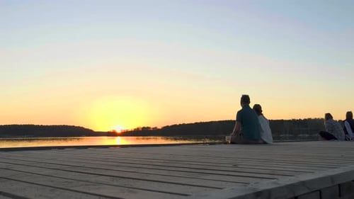 A couple is sitting on a pier by the lake and watching the sunset