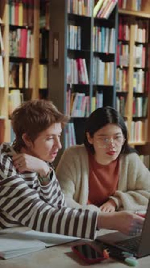 Two Female Group Mates Using Laptop and Talking in Library