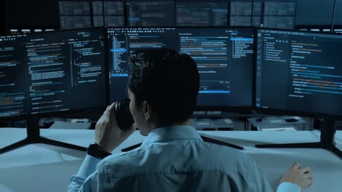 Asiatischer Mann trinkt Kaffee und benutzt Computer, um Code mit mehreren Bildschirmen im Büro zu schreiben