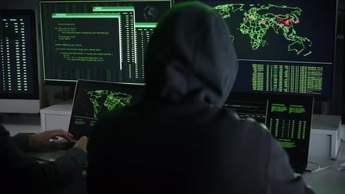 Deux pirates informatiques cagoulés travaillent ensemble et pointent du doigt des écrans d'affichage dans un bureau sombre